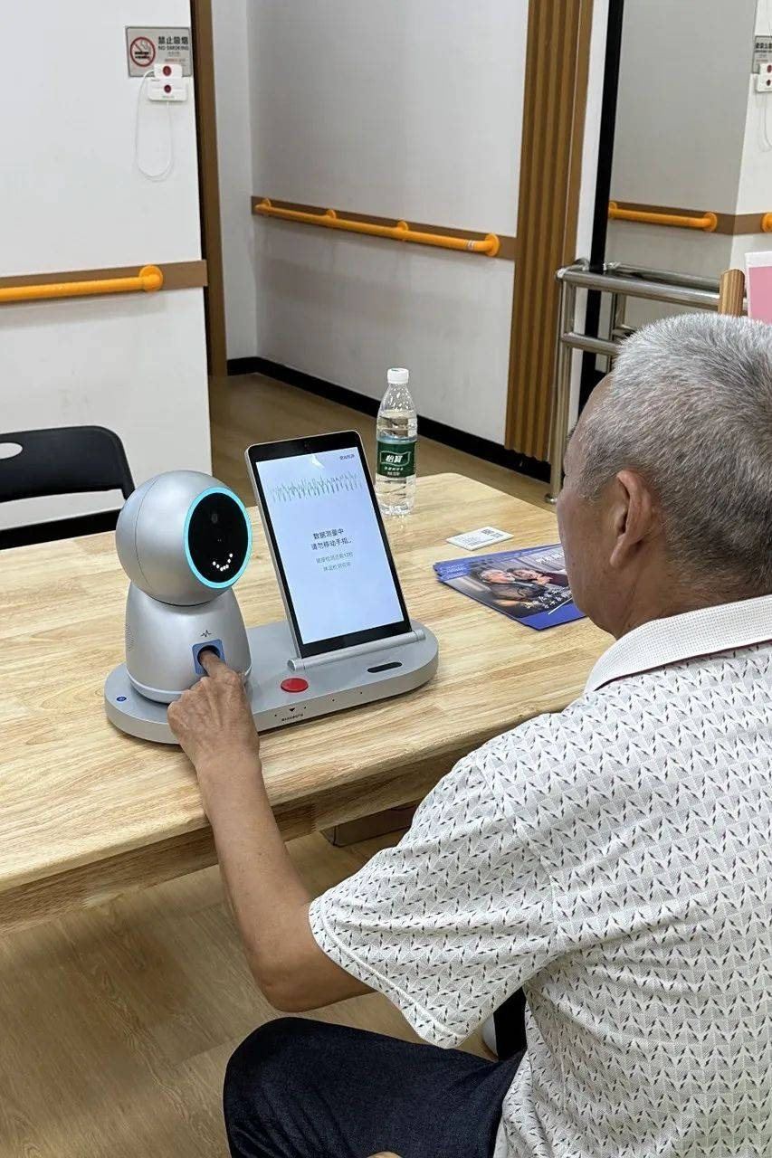 Small desktop companion robot on a table beside an armchair, seen from behind a resident